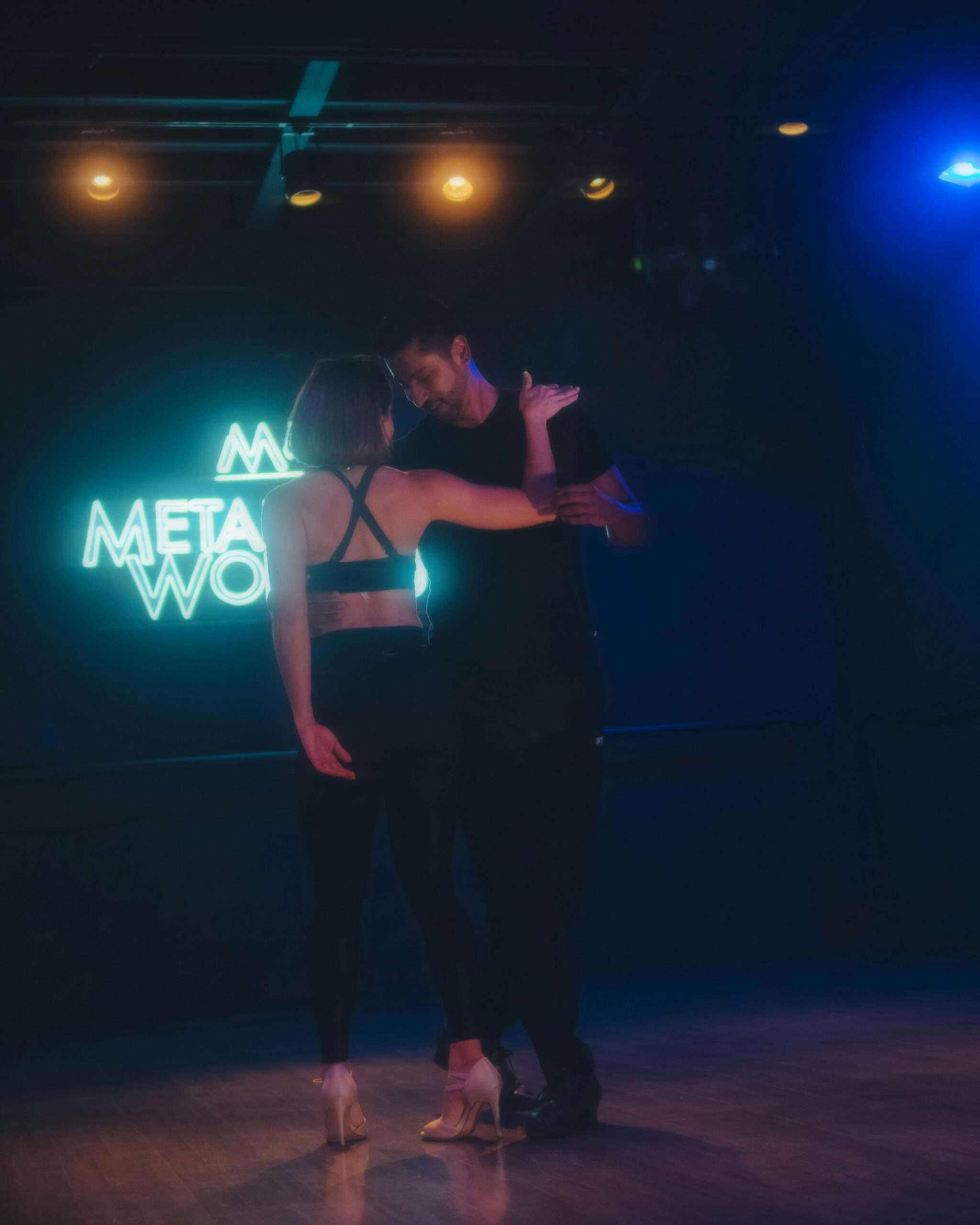 A couple embraces while dancing in a studio with neon lighting and wooden flooring.